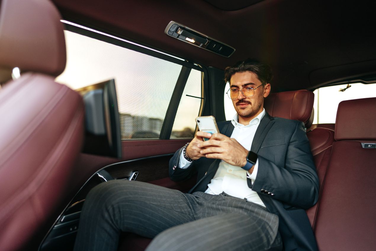 Mann im Anzug, entspannt auf dem Rücksitz einer Luxuslimousine, Smartphone in der Hand – perfekte Vorbereitung für ein Escort Köln Date.